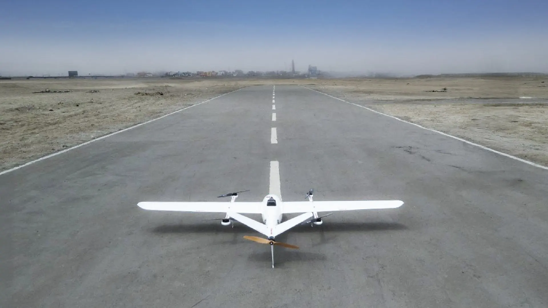 Aeronave de ala fija de Robotic Air Systems sobre una pista en un llano, lista para despegar, con construcciones al fondo entre la bruma